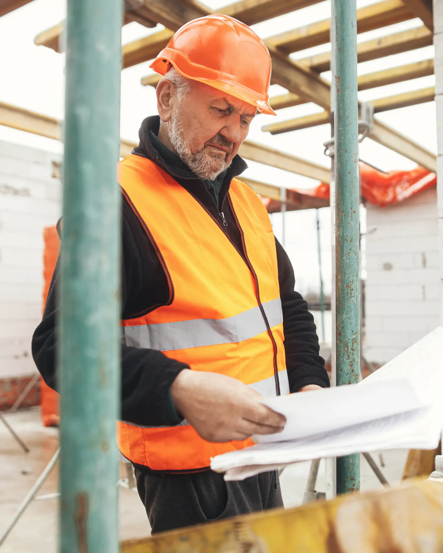 Bauleiter mit orangem Helm und Warnweste liest auf der Baustelle Baupläne, im Hintergrund gestapelte Porenbetonsteine