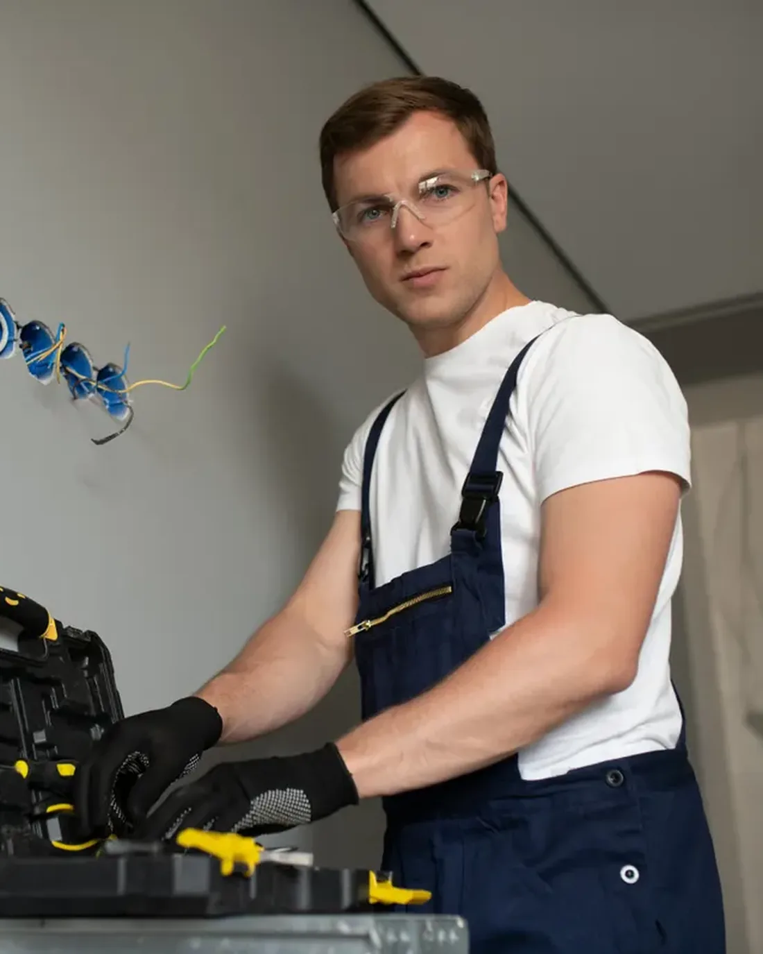 Elektriker mit Schutzbrille und Arbeitshandschuhen greift in einen Werkzeugkoffer, hinter ihm Unterputzdosen mit Leitungen