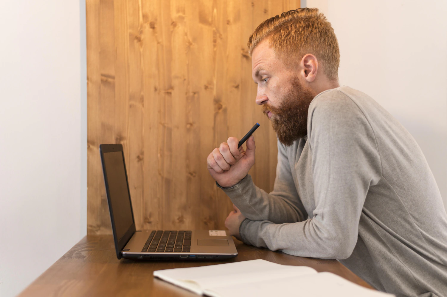 Mann sitzt nachdenklich mit Stift in der Hand vor seinem Laptop und vergleicht Angebote.
